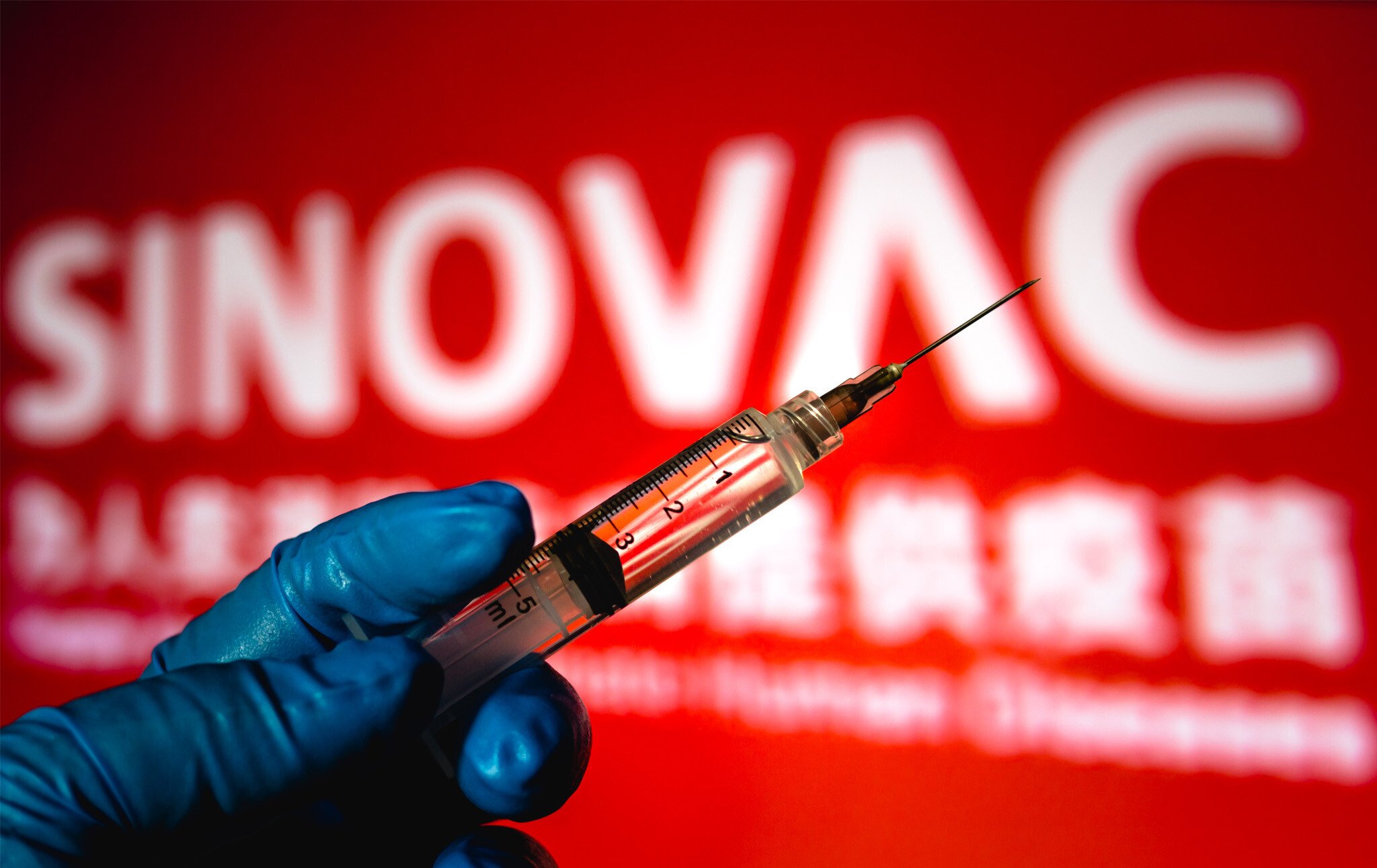 A gloved hand holds a vaccine syringe, in front of a large red sign that says